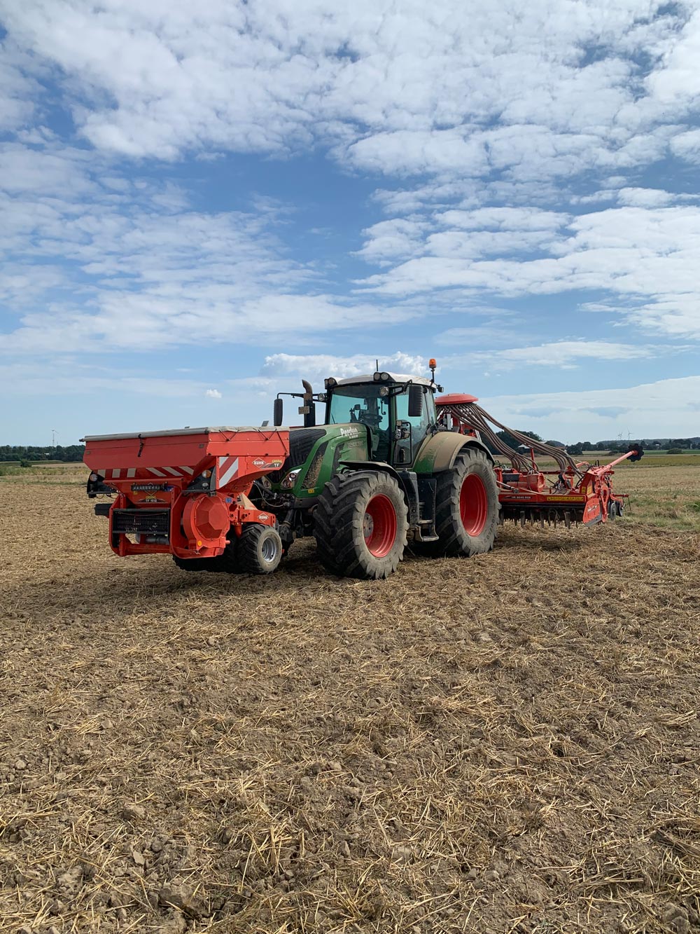 Ein großer grüner Traktor mit roter Landmaschine steht auf einem braunen Ackerfeld